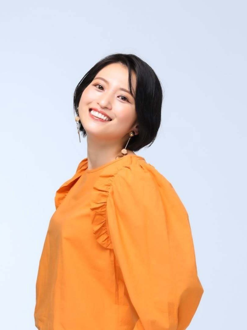 Portrait of a smiling woman with short black hair wearing an orange blouse against a pale blue background.