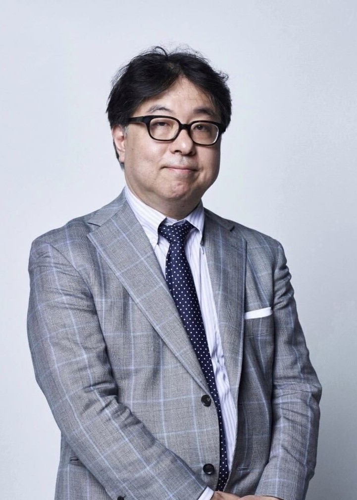 Portrait of a man in a gray plaid suit, white shirt, navy polka-dot tie, and black glasses against a light background.