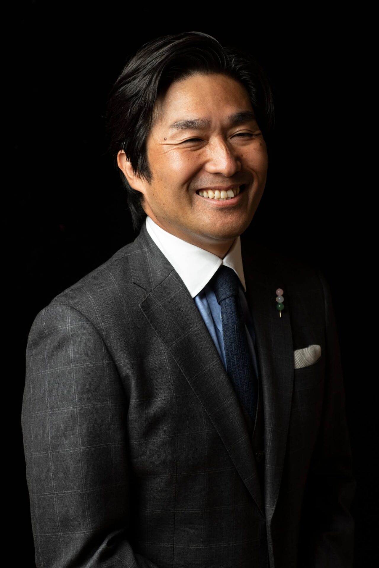 Smiling man in a gray checked suit, white shirt, and blue tie against a black background.