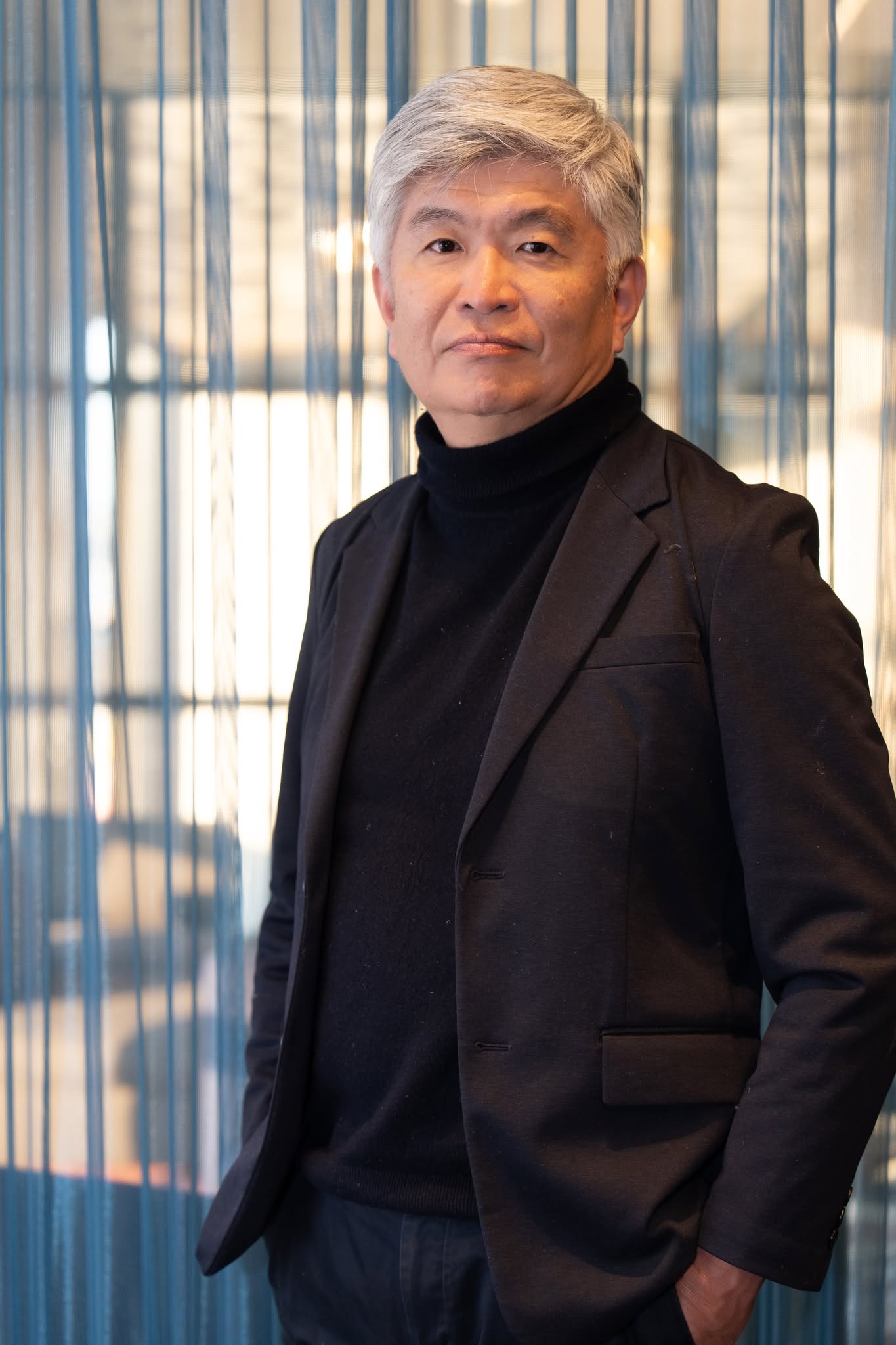Older man with silver hair in a black turtleneck and blazer, standing in front of blue vertical blinds.